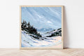 Framed painting of a snowy landscape with trees and mountains on a white wall.