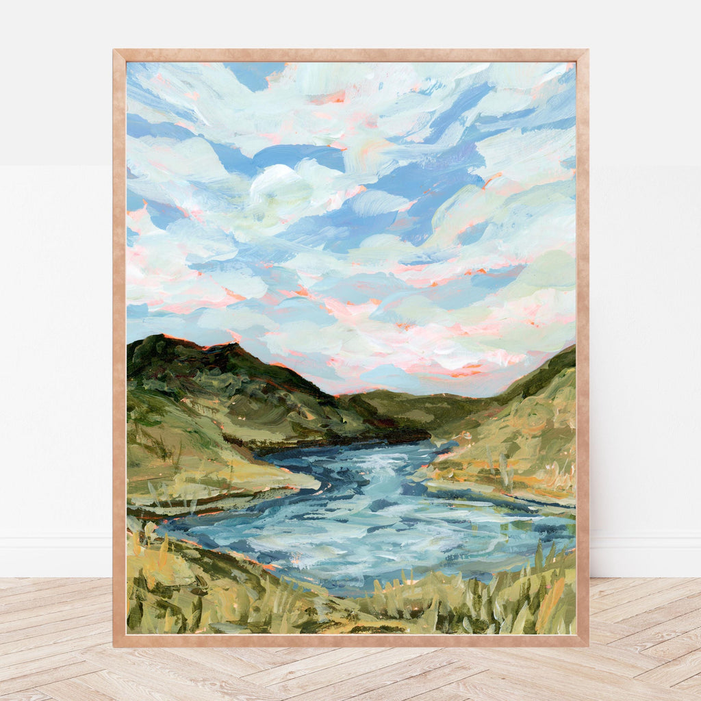 Framed painting of a landscape with a river and green hills on a white wall.