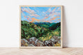 Framed painting of a mountain landscape with a colorful sky on a white wall.