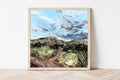 Framed painting of a desert landscape with mountains and clouds on a white wall.