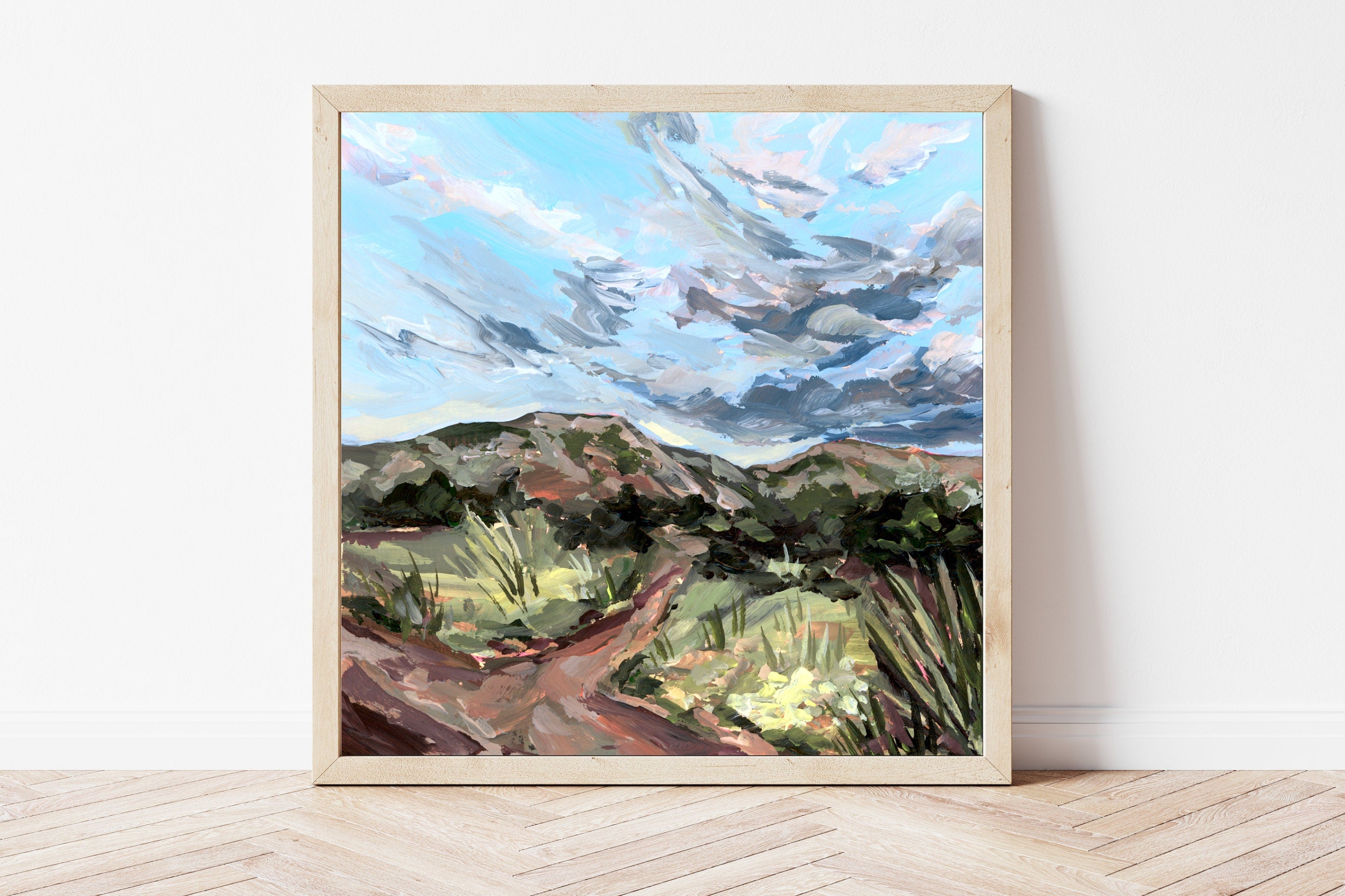 Framed painting of a desert landscape with mountains and clouds on a white wall.
