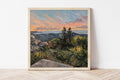 Framed painting of a Maine landscape with trees and a sunset sky on a white wall.