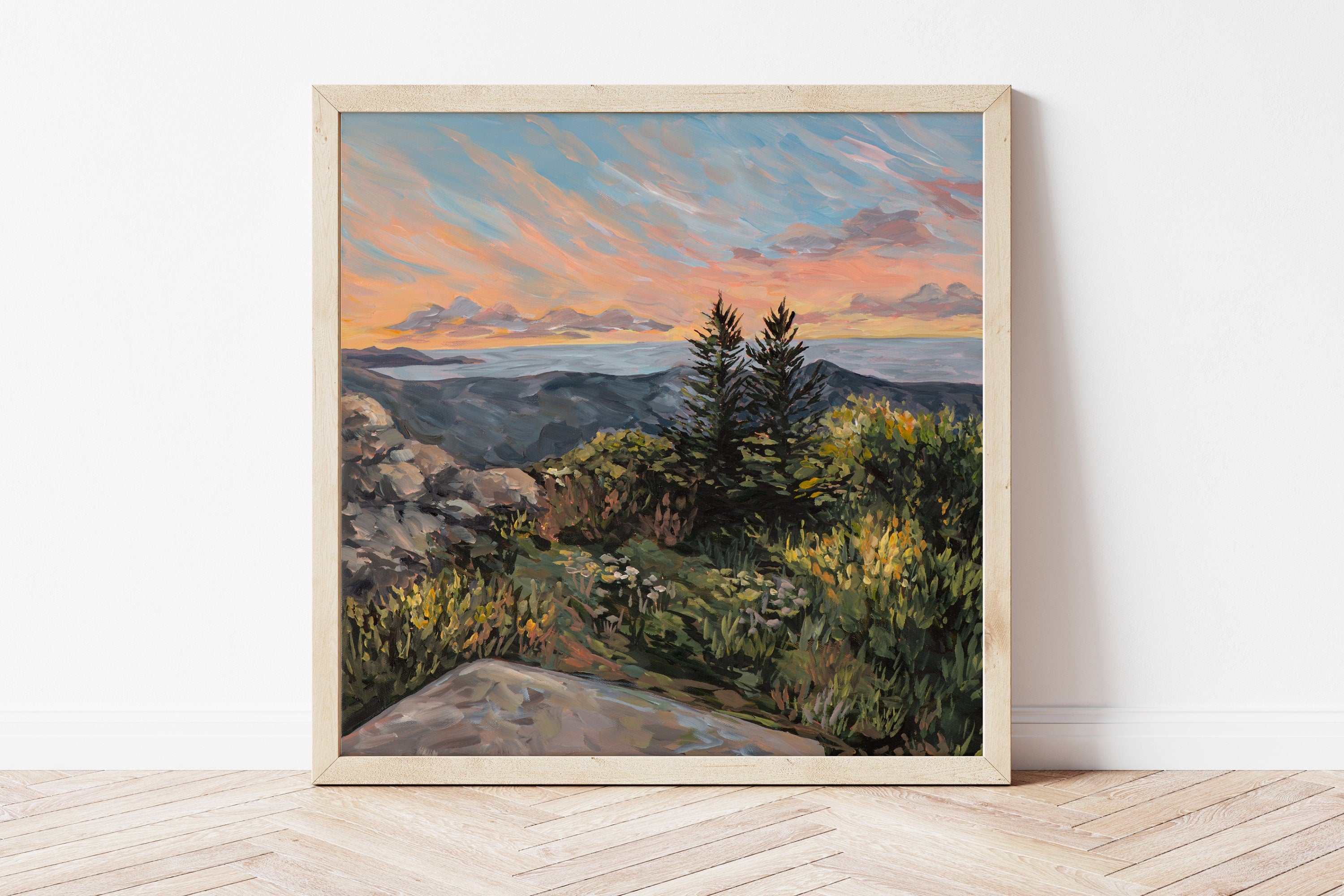 Framed painting of a Maine landscape with trees and a sunset sky on a white wall.