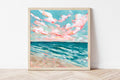 Framed artwork of a beach scene with pink clouds and blue water on a white wall.