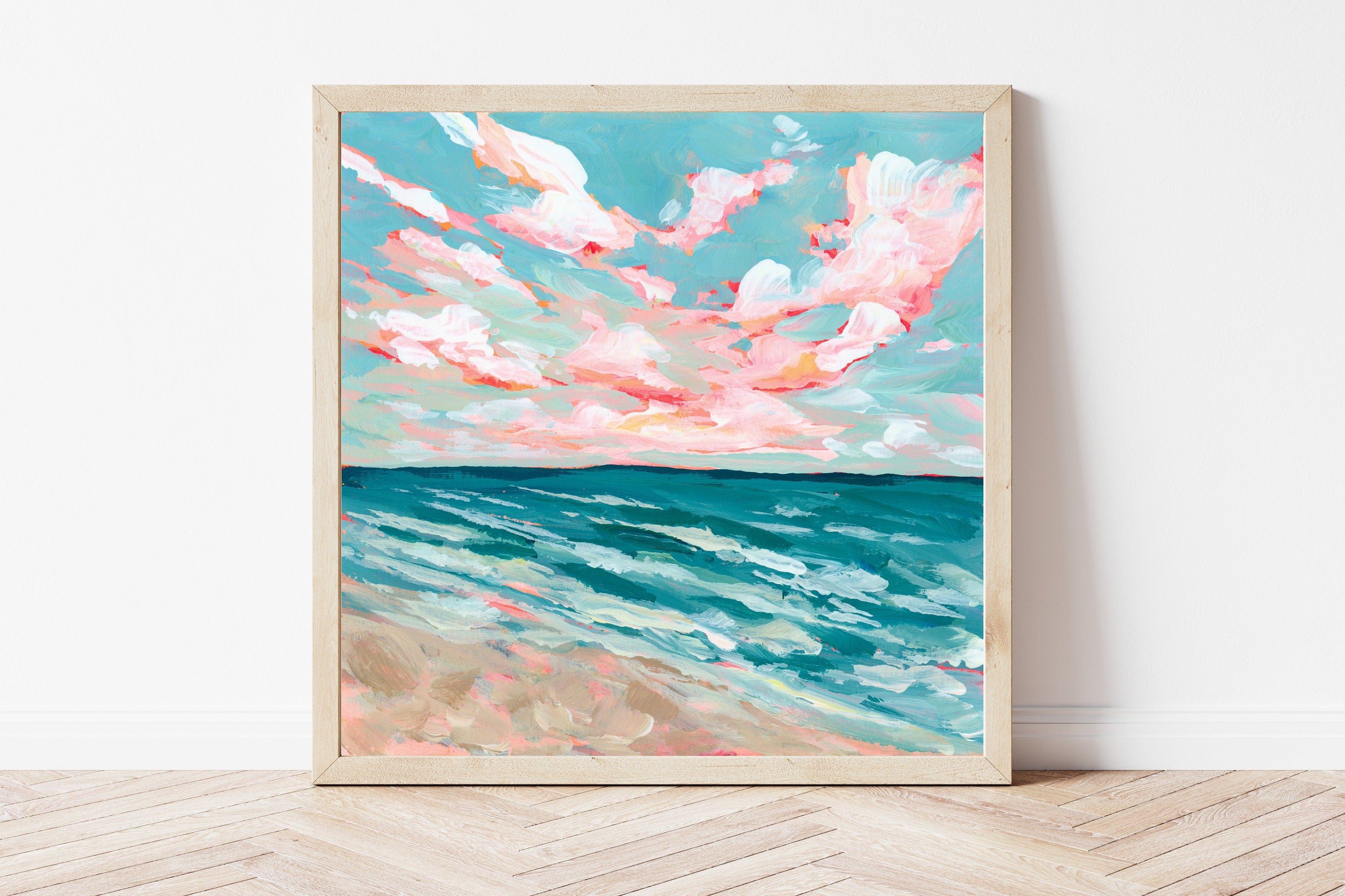 Framed artwork of a beach scene with pink clouds and blue water on a white wall.