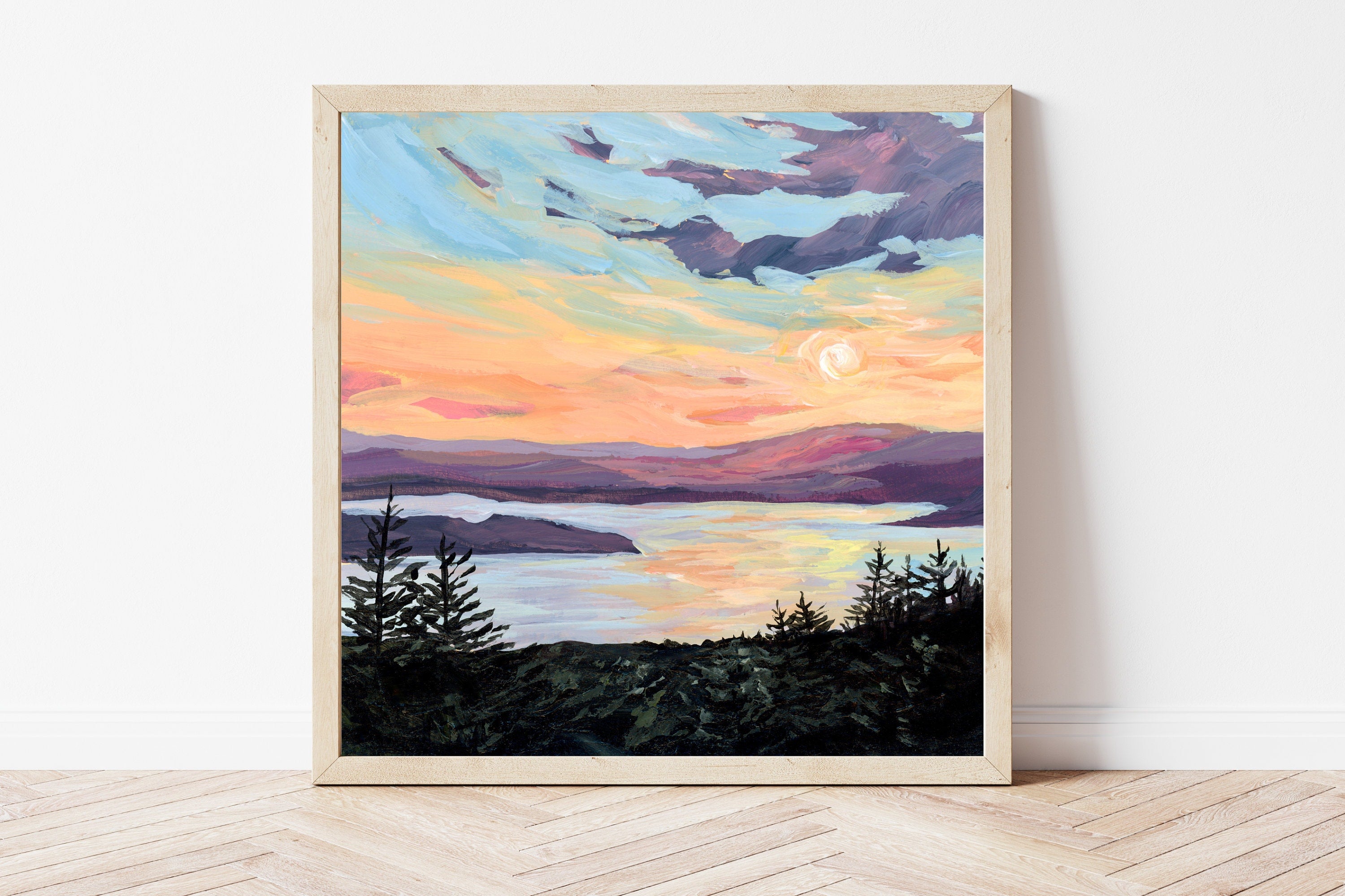 Framed painting of a peaceful Maine sunset over water with the silhouette of trees in the foreground, on a white wall.