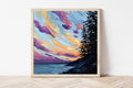 Framed artwork of a colorful Maine sunset over the ocean with trees on a white wall.