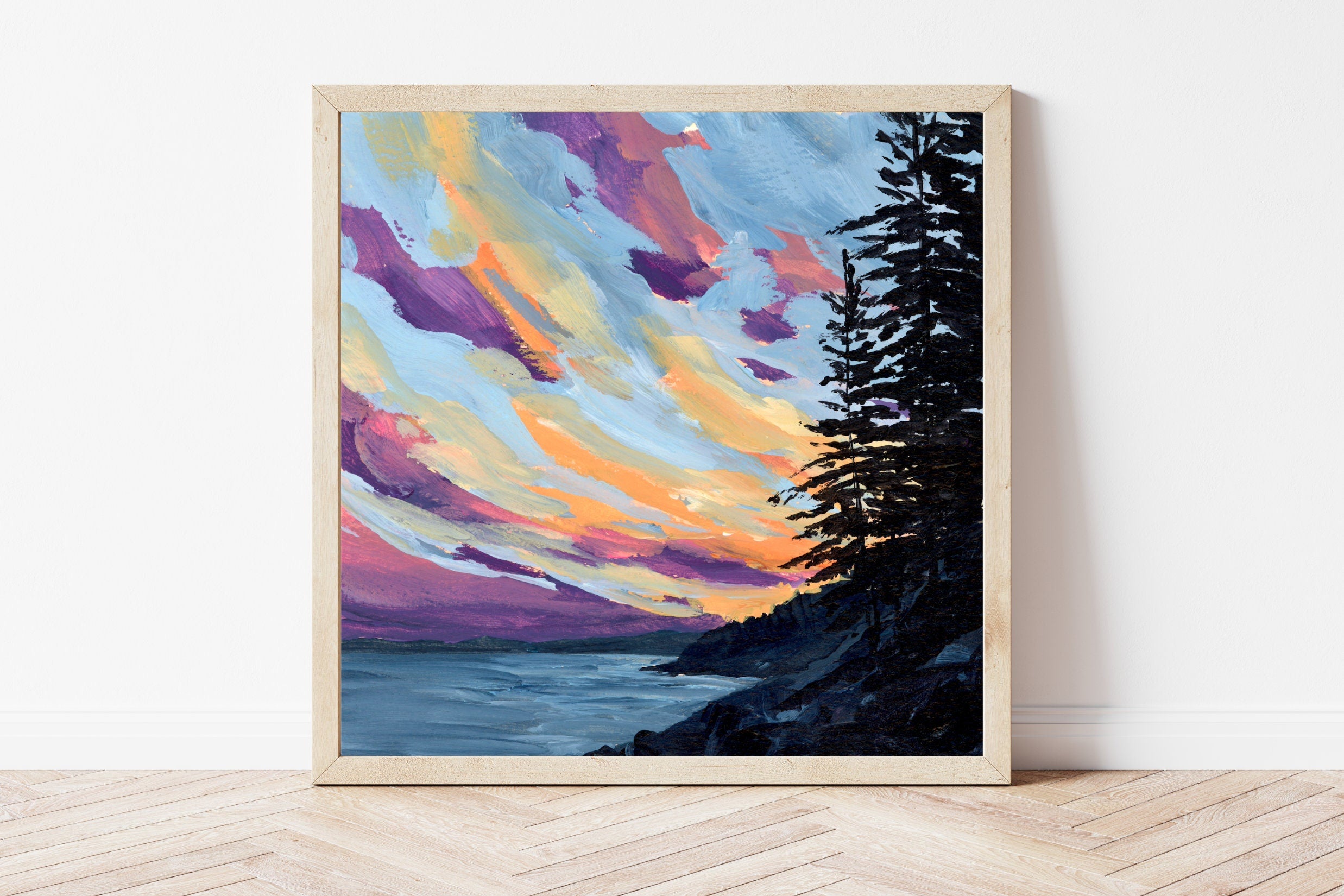 Framed artwork of a colorful Maine sunset over the ocean with trees on a white wall.