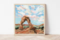 Framed landscape print depicting the landmark "Delicate Arch" located in Arches National Park, Utah. The frame is resting against a white wall. 