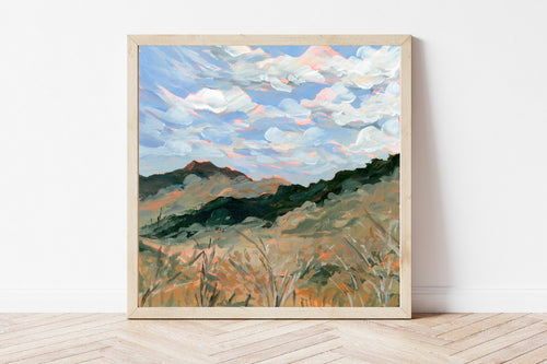 Framed painting of a landscape with mountains and sky on a white wall.