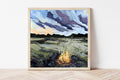 Framed painting of a campfire scene with a landscape in the background on a white wall.