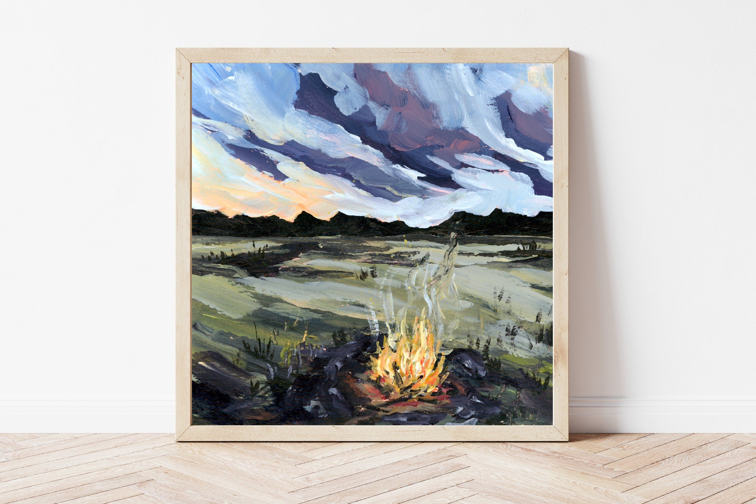 Framed painting of a campfire scene with a landscape in the background on a white wall.