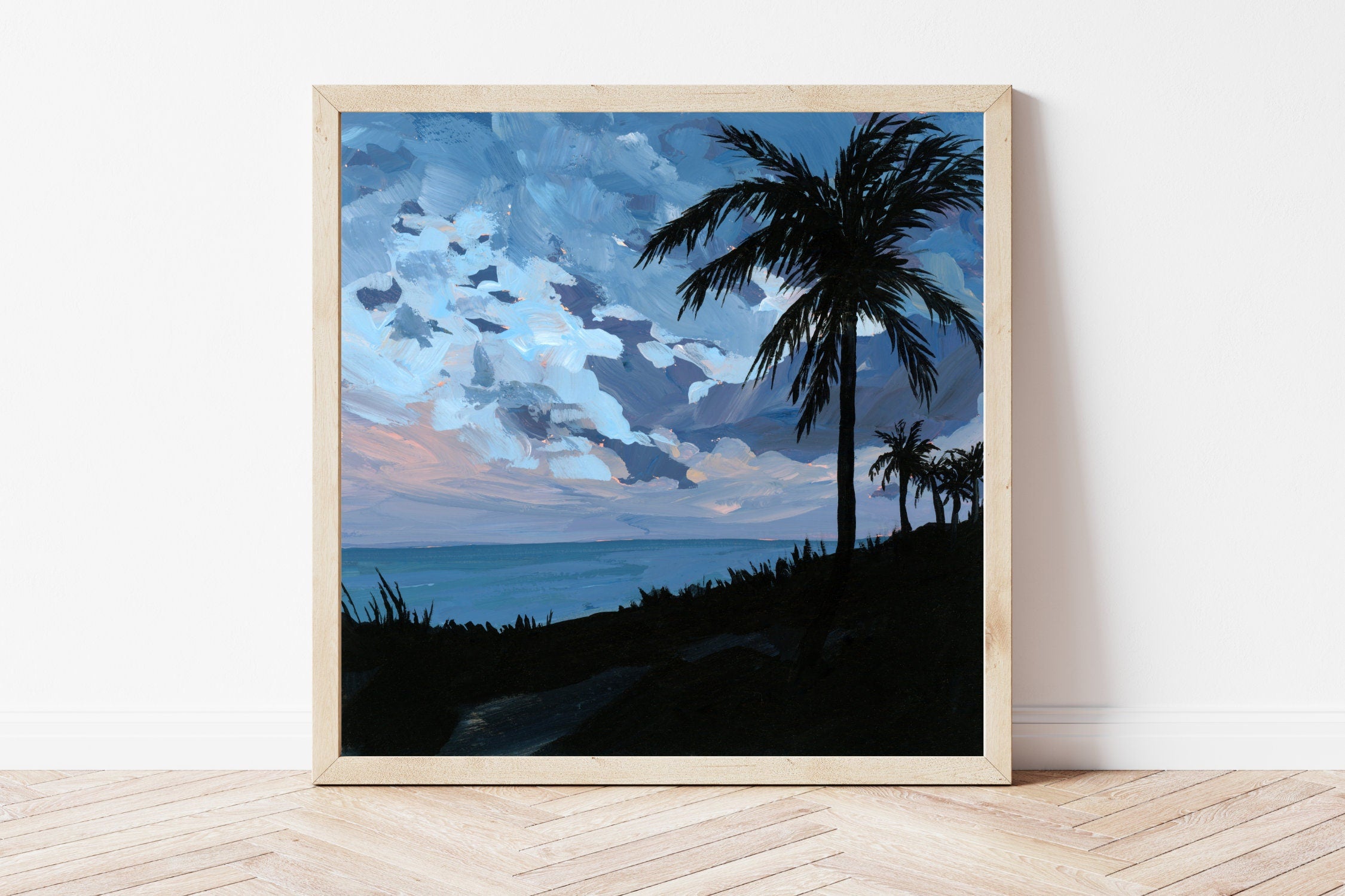 Framed artwork of a tropical landscape at dusk with palm tree silhouettes and ocean on a white wall.