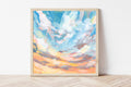 Framed artwork of a colorful sky with clouds on a white wall.