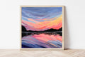 Framed artwork of a colorful sunset over water on a white wall.