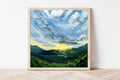Framed painting of a sun rising over mountains on a white wall.