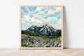 Framed painting of a mountain landscape with flowers on a white wall.
