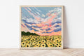 Framed artwork of a sunflower field with a colorful sky on a white wall.