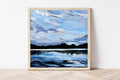 Framed painting of a landscape with a blue sky and water on a white wall.