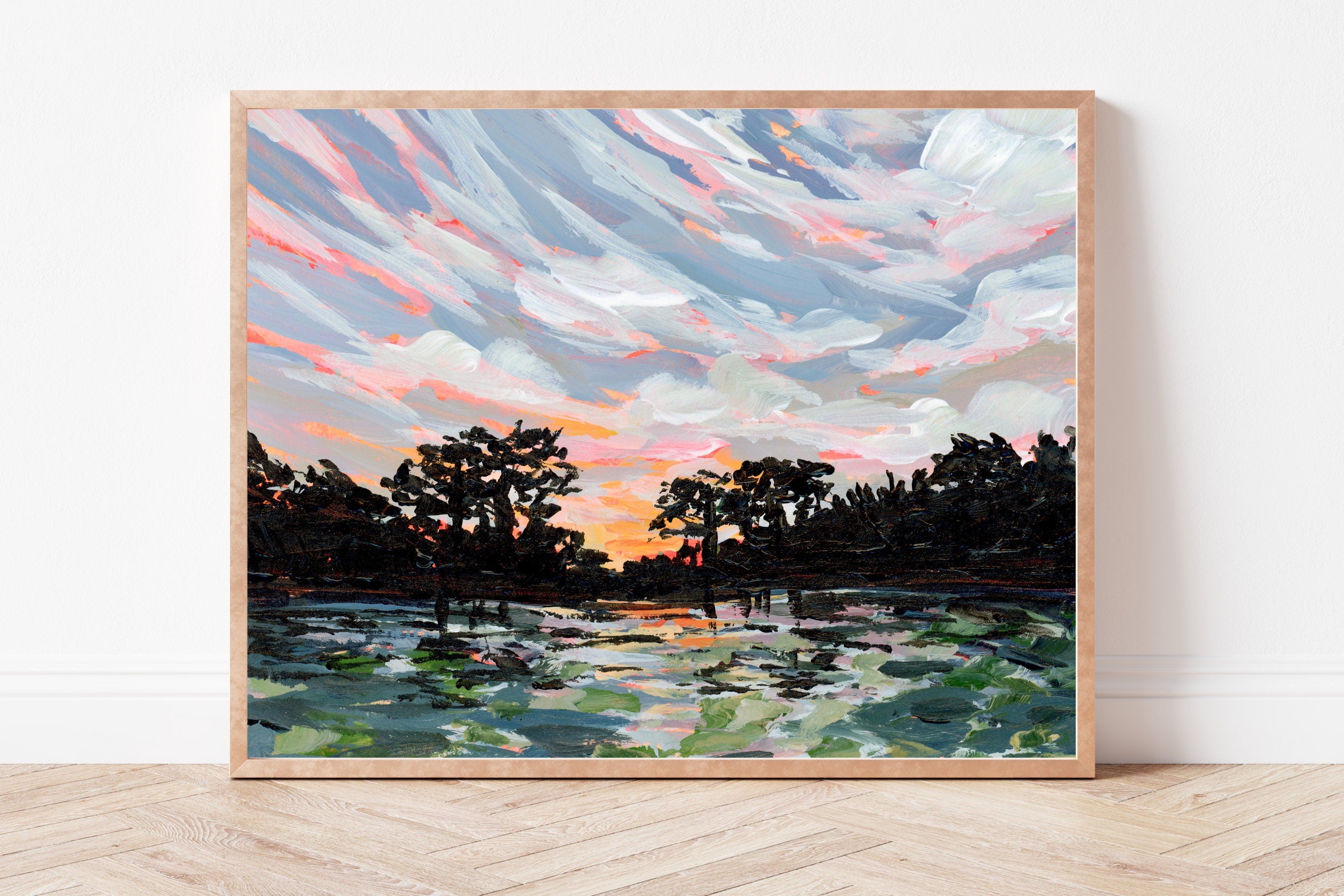 Framed painting of a sunset over water with trees on a white wall.