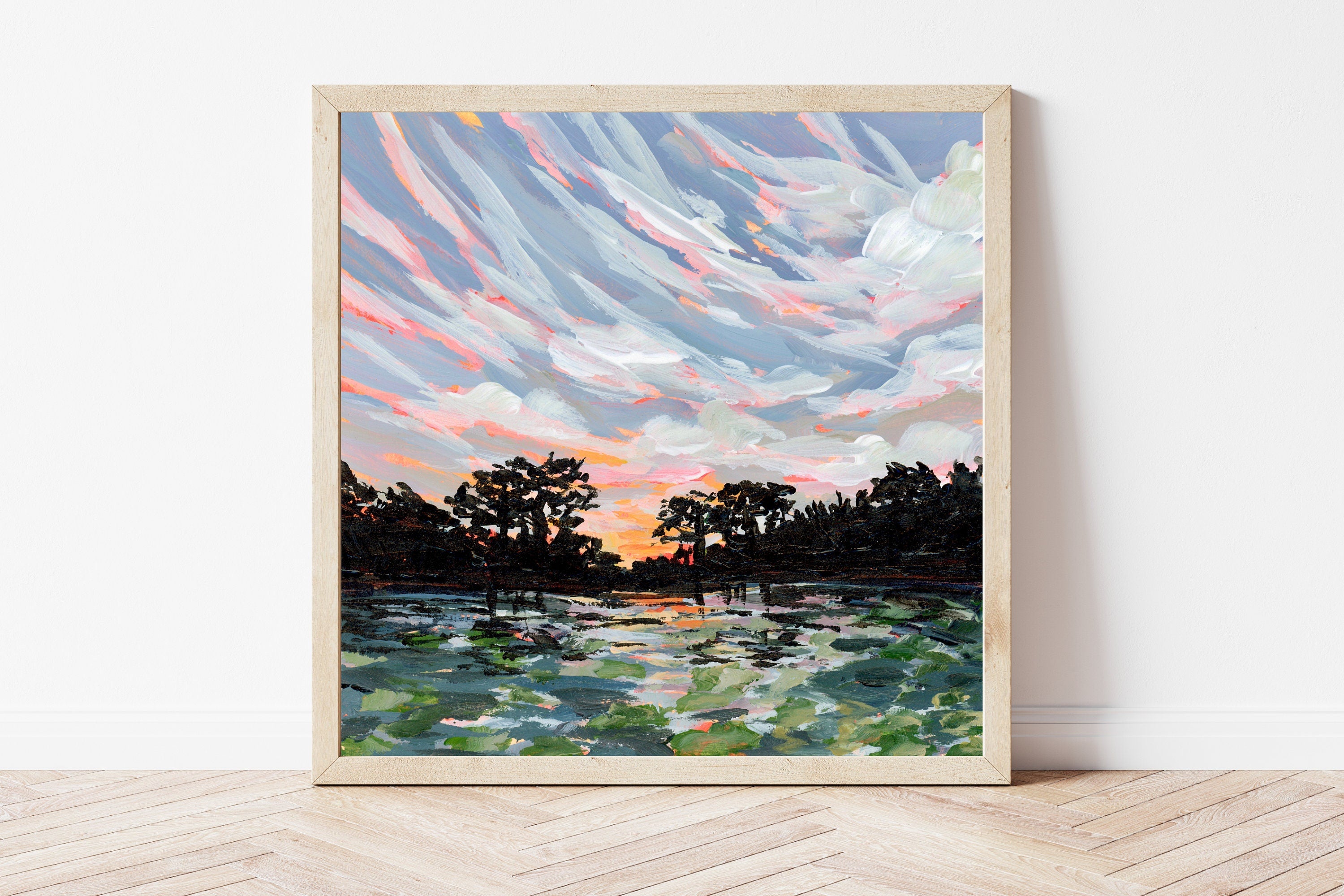 Framed painting of a sunset over water with trees, on a white wall.