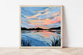 Framed painting of a sunset over water with a wooden frame on a white wall.