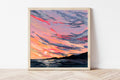Framed artwork of a colorful sunset over water on a white wall.