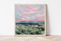 Framed painting of a rural Wyoming landscape with pink clouds and mountains on a white wall.