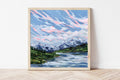 Framed print of an Alaskan mountain landscape with a lake and clouds on a white wall.