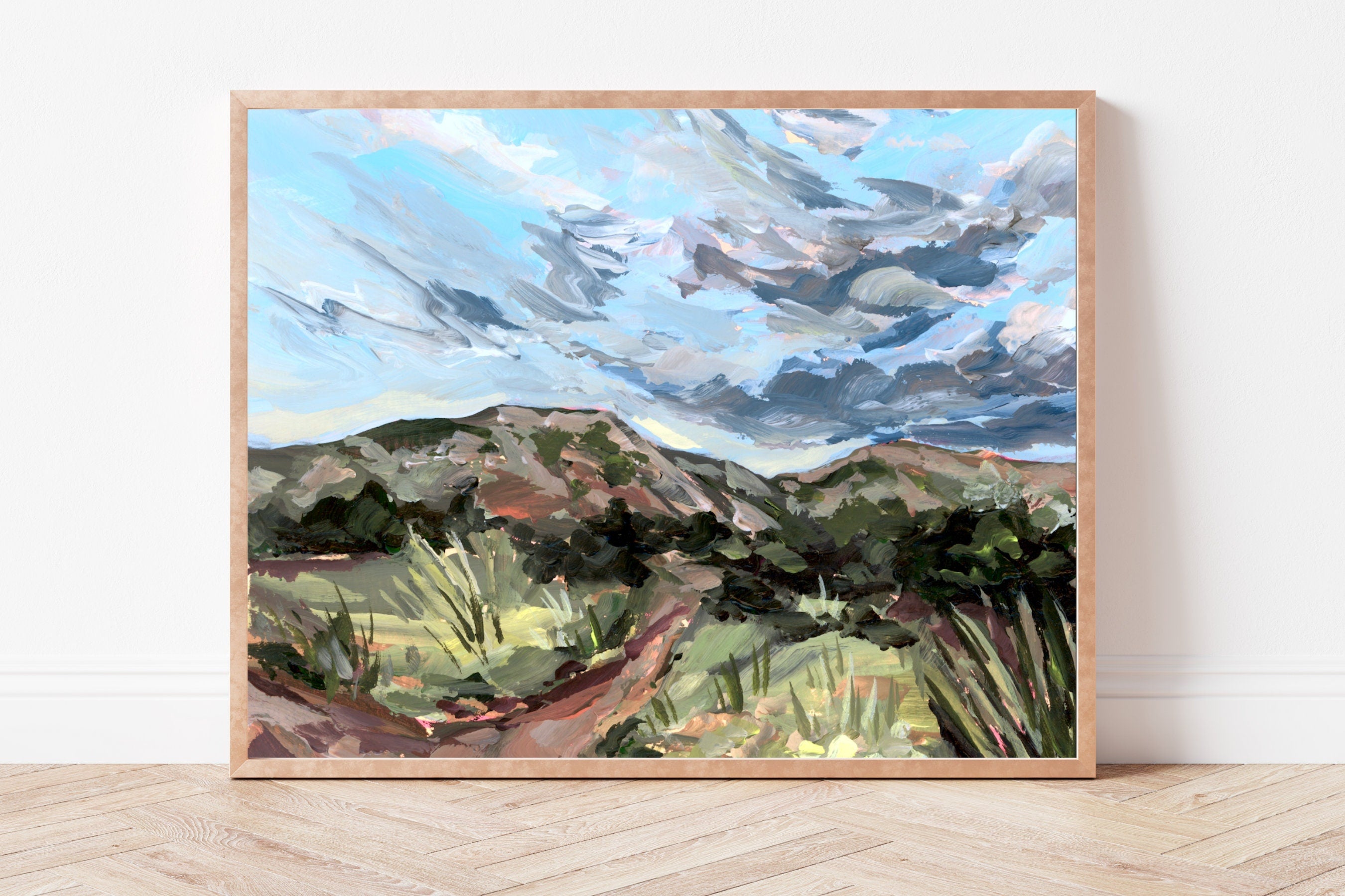 Framed painting of a desert landscape with mountains and sky on a white wall.