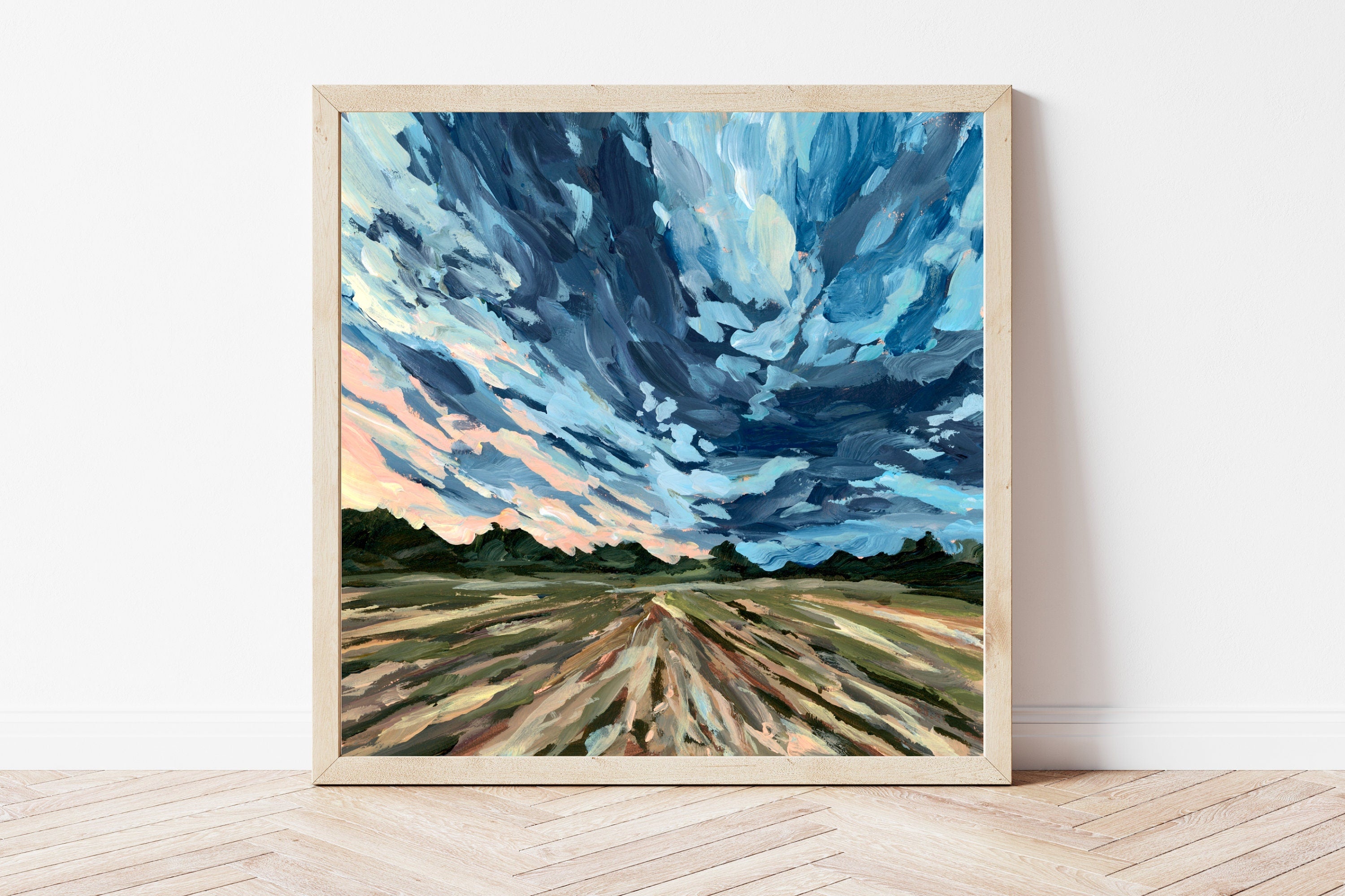 Framed art print of a painting featuring bold storm clouds over a rural landscape. The frame is sitting on a wooden floor, propped against a white wall. 