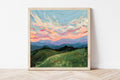 Framed painting of a mountain landscape with a colorful sky on a white wall.